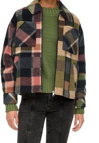 Chaqueta de forro polar de cuadros en tonos verde, beige, rosa y negro; cuenta con dos bolsillos frontales y un cuello con solapas. Llevada sobre un suéter verde.