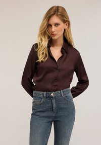 Blonde vrouw met gouden oorbellen, een donkerbruine satijnen blouse met knopen en hoge, blauwe jeans tegen een eenvoudige achtergrond.