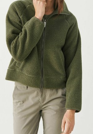 Personne portant une veste polaire zippée vert olive et un pantalon beige clair, une main près du menton et l'autre pendant.