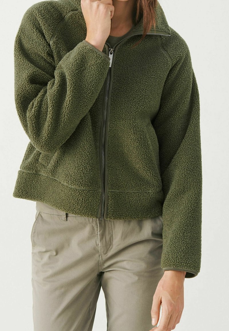 Personne portant une veste polaire zippée vert olive et un pantalon beige clair, une main près du menton et l'autre pendant.