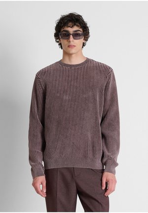 Giovane uomo con occhiali da sole scuri, maglione viola a coste a maniche lunghe e pantaloni viola abbinati su uno sfondo chiaro uniforme.