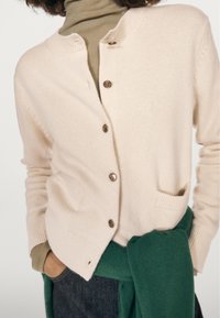 Light beige cardigan with button front, ribbed cuffs and pockets. Worn over a green sweater and a fitted olive turtleneck. Soft texture.
