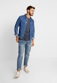Jeansjakke i mellomblått, med knappestolpe foran, brystlomme og rullede ermer. Kombinert med en grå t-skjorte og lyseblå jeans.