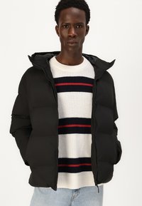 Sort pufferjakke med hætte, quiltet design, frontlynlås, båret over en hvid sweater med marineblå og røde vandrette striber.