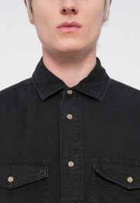 Black denim shirt with a collar, featuring two front pockets and beige buttons. The fabric has a smooth texture with a slight sheen.