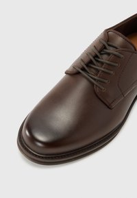 Chaussure en cuir marron avec un bout arrondi, fermeture à lacets et une finition légèrement brillante. Présente des coutures renforcées le long de la semelle.
