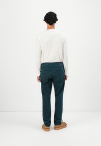 Homme se tenant de dos, portant une chemise blanche à manches longues, un jean vert foncé et des chaussures marron clair, devant un fond blanc uni.