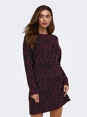 Femme portant une robe en maille à manches longues imprimée léopard rouge foncé et noire, posant avec une main sur le côté et regardant vers la gauche.