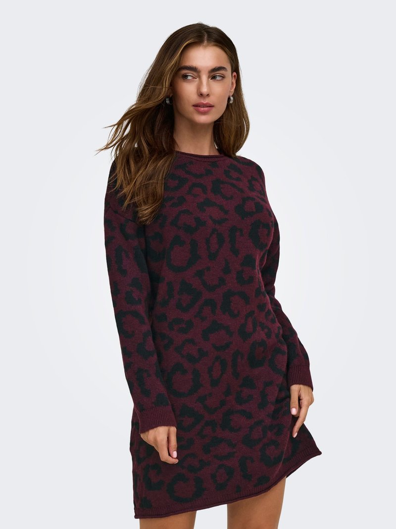 Femme portant une robe en maille à manches longues imprimée léopard rouge foncé et noire, posant avec une main sur le côté et regardant vers la gauche.