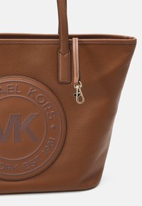 Borsa tote in pelle marrone con un logo circolare impresso, superficie texturizzata e doppi manici. Include un dettaglio in metallo dorato con chiusura.