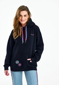 Naine, kes kannab musta kapuutsiga pusa, millel on roosad ja sinised nöörid, ripsmembroidery, lillemustriga plaastrid ning helesinised teksad roosade küljeribadega.