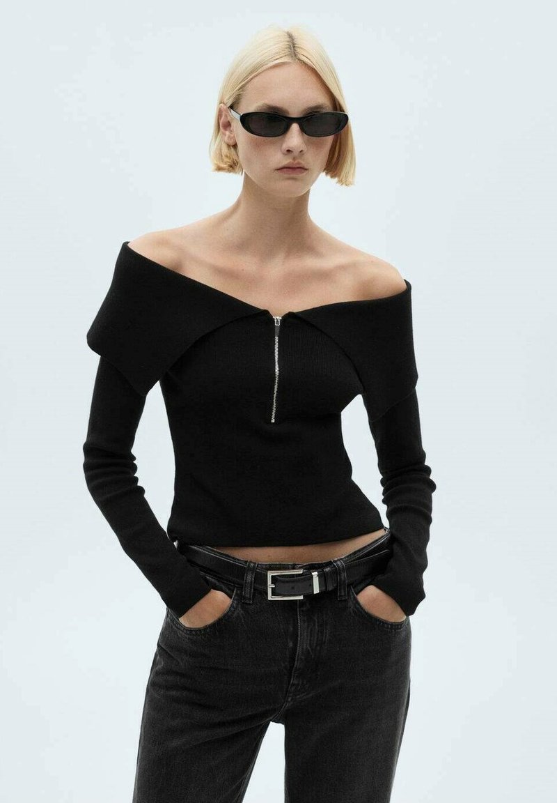 Zwarte gebreide off-shoulder top met een rits in het midden, gecombineerd met high-waisted donkere jeans en een zwarte riem met een zilveren gesp.