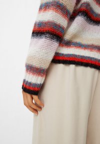 Sweter z dzianiny w kolorowe poziome paski w odcieniach czerwieni, niebieskiego i kremowego, z puszystą teksturą i ściągaczami. W tle miękka beżowa tkanina.