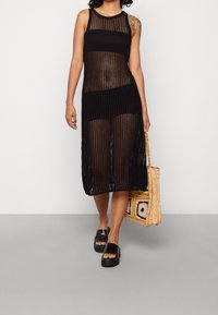 Abito nero a maglia senza maniche con una vestibilità ampia e tessuto trasparente, caratterizzato da un orlo dritto. Abbinato a sandali neri con plateau e una borsa di paglia.