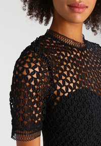 Černý háčkovaný top s krátkými rukávy, složitým smyčkovým vzorem a pevnou černou podšívkou s texturovaným povrchem.