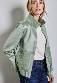 Giacca verde menta con colletto in pelliccia sintetica e tasche a toppa, caratterizzata da una texture esterna liscia e cordini regolabili sull'orlo.