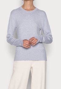 Pull gris clair à manches longues, encolure ronde, tissu maille douce, poignets et ourlet côtelés, porté avec un pantalon large couleur crème.