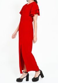 Rochie maxi roșie cu mâneci scurte cu volane, despicătură în față și talie elastică. Asortată cu pantofi cu platformă negri. Textură netedă, design elegant.