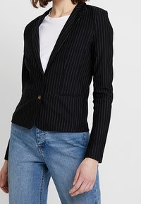 Černý blazer s bílými vertikálními pruhy, se zapínáním na jeden knoflík, klopami a dvěma předními kapsami, v kombinaci s modrými džíny.