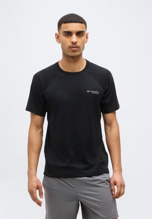 Joven con una camiseta negra de manga corta Columbia Titanium y pantalones cortos grises, de pie frente a un fondo claro y liso.