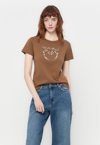 T-shirt marron à manches courtes avec un design orné de motifs métalliques à l'avant, associé à un jean en denim bleu clair à coupe décontractée.