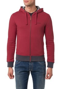 Felpa rossa con zip e cappuccio regolabile, polsini e orlo grigi, tasche frontali e fodera interna a fantasia. Modello che indossa jeans blu.