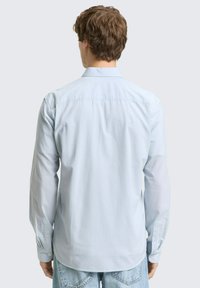 Man die wegkijkt, draagt een lichtblauw overhemd met lange mouwen en lichtgewassen jeans voor een effen grijze achtergrond.