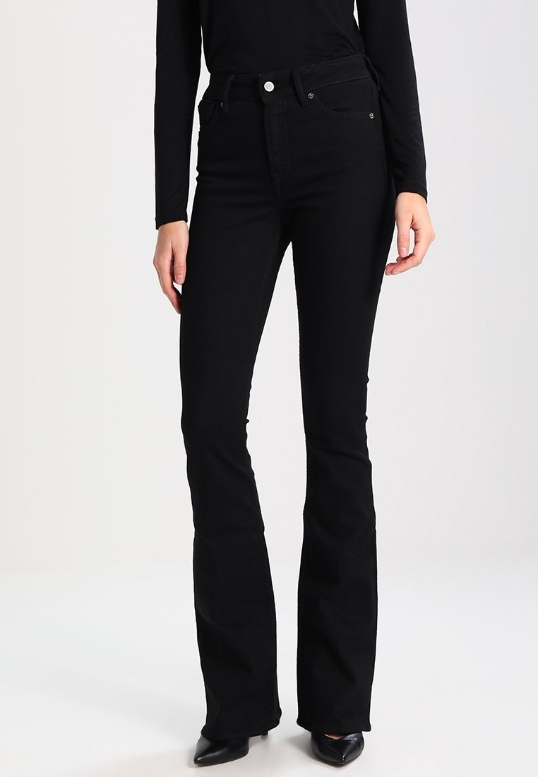 Kobieta ubrana w czarne jeansy z wysokim stanem i rozszerzanymi nogawkami oraz czarne buty z zakrzywionym noskiem, stojąca na tle jasnego, jednolitego tła.