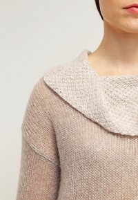 Pull en tricot beige clair avec un large col replié. La texture est douce, présentant un mélange de fils fins et épais pour plus de profondeur.