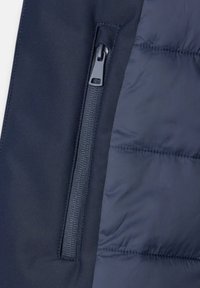 Sezione giacca puffer blu navy con tasca con zip e hardware nero, tessuto morbido con leggero splendore e texture trapuntata.