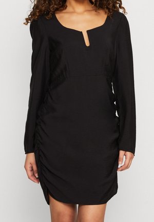 Vestido negro de manga larga con escote cuadrado en muesca y lados fruncidos, llevado por una persona con cabello rizado visible desde los hombros hacia abajo.
