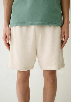 Person iført beige knælange shorts og en grøn t-shirt med hænder afslappet langs siderne mod en ensfarvet baggrund.