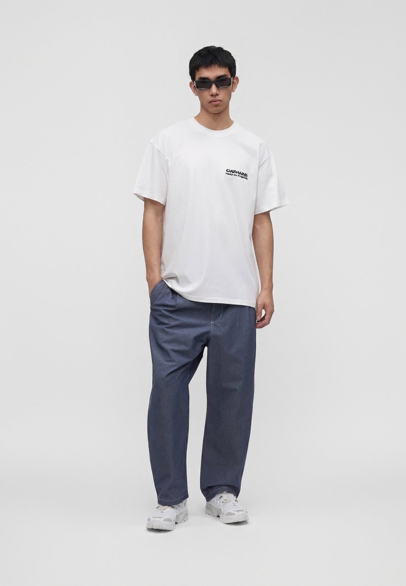 Giovane uomo con occhiali da sole neri, t-shirt bianca Carhartt, pantaloni blu larghi e sneakers bianche, in piedi davanti a uno sfondo grigio uniforme.