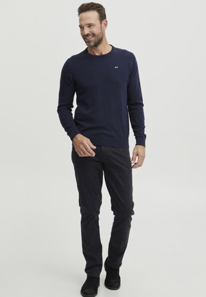 Marineblauer Strickpullover mit rundem Halsausschnitt, langen Ärmeln und Logo-Detail auf der Brust, kombiniert mit schwarzen Slim-Fit-Hosen und schwarzen Stiefeln.