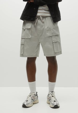 Homme portant un short cargo gris clair avec plusieurs poches, des chaussettes blanches montantes et des baskets argentées, debout avec les jambes visibles.