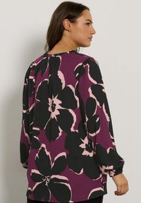 Blusa viola con grande motivo floreale nero e rosa. Presenta un colletto rotondo, maniche lunghe con polsini elasticizzati e tessuto testurizzato.