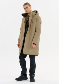 Beige lange jas met een capuchon en een voorrits, zwarte binnenvoering, gecombineerd met zwarte cargobroek en veterschoenen. Bevat verstelbare manchetten.