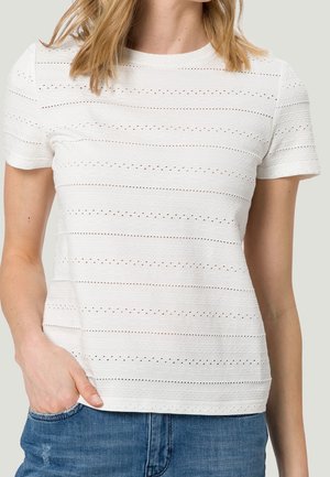 Femme portant un haut blanc texturé à manches courtes avec des bandes perforées horizontales, jean bleu, main dans la poche, fond neutre.