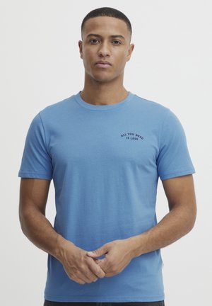 Lyseblå bomulds t-shirt med korte ærmer og rund halsudskæring, prydet med sort tekst "ALL YOU NEED IS LESS" på venstre brystområde.