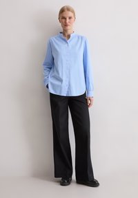 Kobieta stojąca na tle jasnej ściany, ubrana w jasnoniebieską koszulę na guziki, czarne spodnie z szerokimi nogawkami i czarne buty, z rękami w kieszeniach.