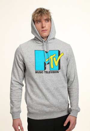 Uomo con felpa grigia con cappuccio su, con grande logo MTV Music Television in blu e giallo, cappuccio alzato, che guarda leggermente di lato.