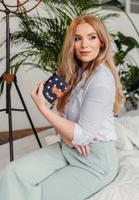 Black wallet with white polka dots and orange accents, featuring a zip closure. Model wearing a light blue blouse and green pants, seated.