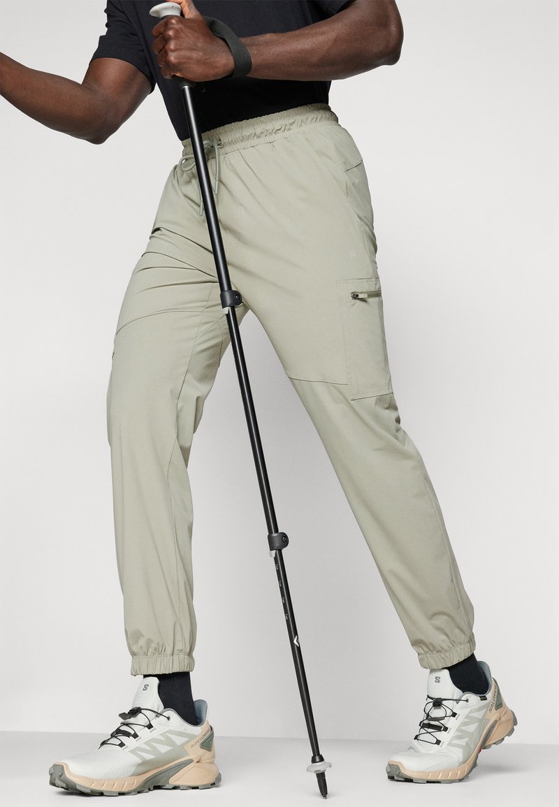 Pantaloni elasticizzati grigio chiaro con vita elastica e tasche laterali, dal taglio affusolato. Abbinati a scarpe da trekking bianche e grigie per attività all'aperto.