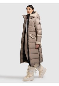 Geacă lungă bej matlasată, cu glugă căptușită cu blană, fermoare negre și buzunare laterale. Purtați peste o ținută mulată și asortați cu cizme albe.