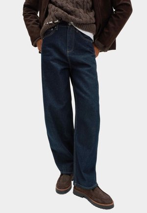 Osoba ubrana w ciemnoniebieskie szerokie dżinsy, brązowe zamszowe buty, brązowy sweter w warkocze i ciemnobrązową kurtkę z rękami w kieszeniach.