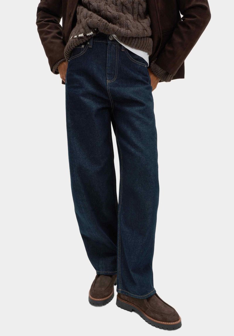 Persona che indossa jeans a gamba larga blu scuro, scarpe in camoscio marroni, un maglione marrone a treccia e una giacca marrone scuro con le mani nelle tasche.