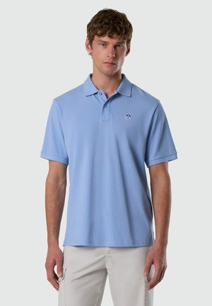 Uomo con capelli biondi e ricci, che indossa una polo azzurro chiaro e pantaloni beige, in piedi davanti a uno sfondo grigio chiaro uniforme.