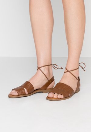 Sandales plates en cuir tressé marron avec des lanières à enrouler autour de la cheville, portées pieds nus sur un fond clair uni.