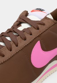 Hnědé tenisky s růžovým Nike swoosh, texturovaná tkanina, krémové tkaničky a polstrovaný jazyk. Podešev má růžové akcenty po celé základně.