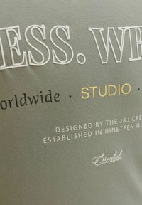 Γκρι ύφασμα με υφασμένο λευκό και χρυσό κείμενο. Κύριες φράσεις περιλαμβάνουν τα «WORLDWIDE», «STUDIO» και «ESSENTIALS». Απλός και μοντέρνος σχεδιασμός.